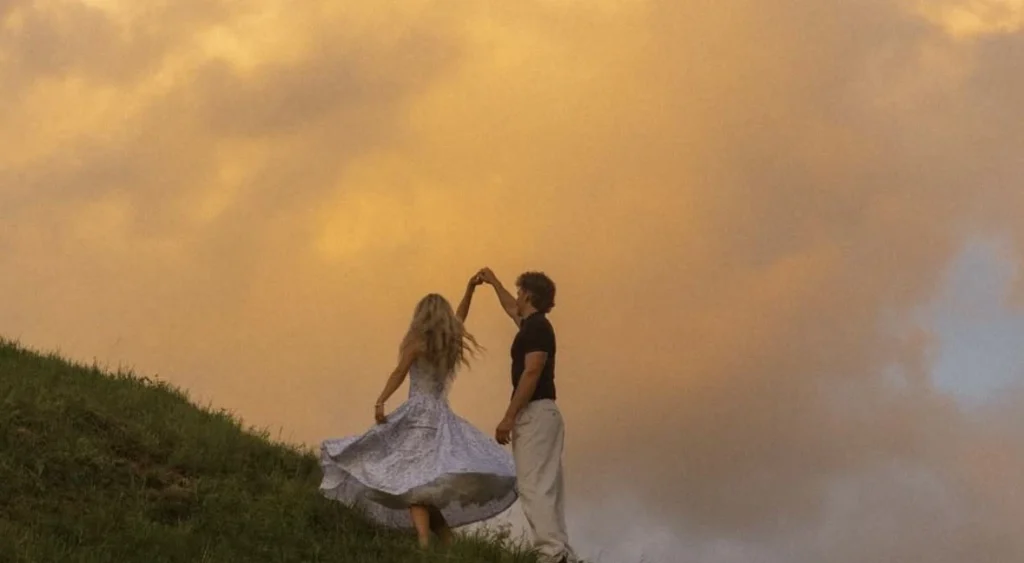 A couple dances on a grassy hill at sunset. The woman in a flowing dress twirls elegantly, while the sky glows with soft golden hues, evoking a romantic, serene mood.