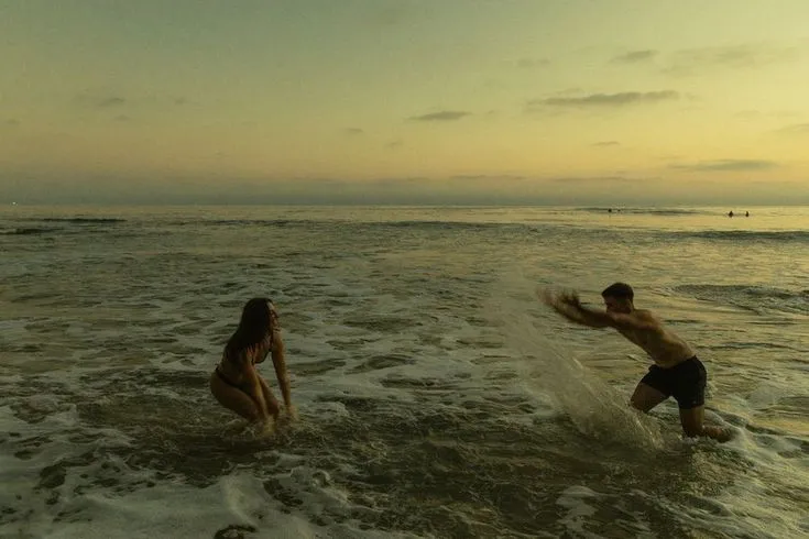 Two people playfully splash water at each other in the ocean at sunset. The sky is a warm blend of yellows and oranges, creating a joyful and relaxed atmosphere.