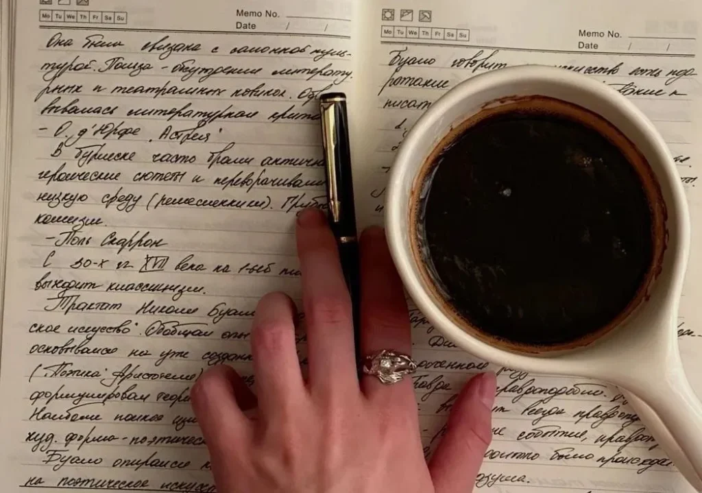 A hand with a pen rests on a handwritten notebook with cursive text, next to a cup of coffee. The ambiance is warm and studious.